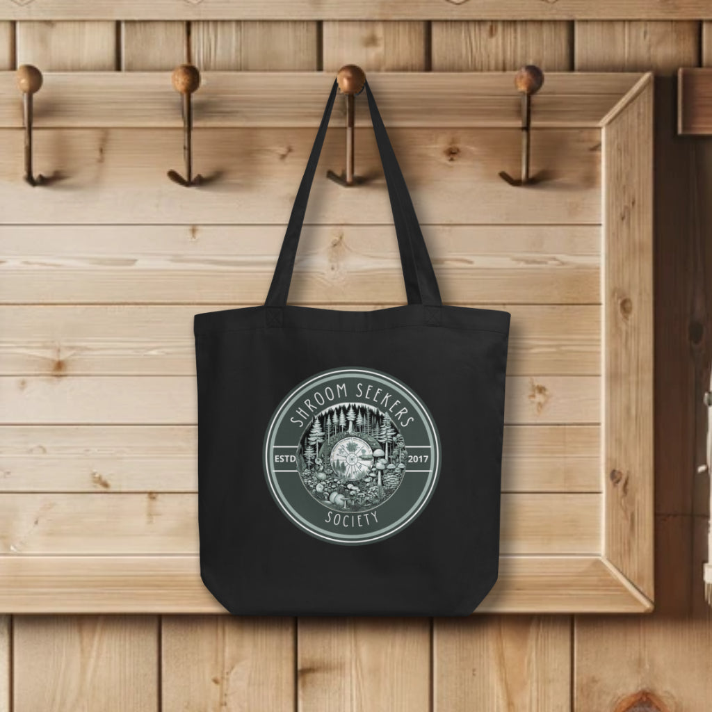 Display of 'Shroom Seekers Society' Tote Bag in black, hanging on wall hooks, depicting a vibrant forest ecosystem with mushrooms and pine trees in a circular pattern, symbolizing the unity and exploration of nature's hidden gems.
