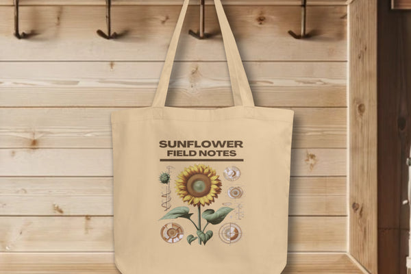 Display of 'Sunflower Field Notes' Tote Bags in oyster, hanging on a wall hook, adorned with a detailed botanical illustration of a sunflower, showcasing the blend of artistry and sustainability.