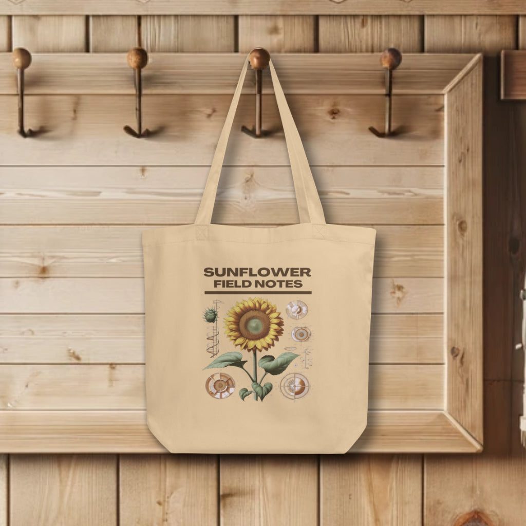 Display of 'Sunflower Field Notes' Tote Bags in oyster, hanging on a wall hook, adorned with a detailed botanical illustration of a sunflower, showcasing the blend of artistry and sustainability.